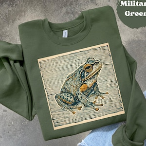 May include: A military green sweatshirt featuring a detailed illustration of a frog within a rectangular frame. The frog design incorporates intricate patterns in shades of teal, brown, and gold. The text "Military Green" is in a circle in the upper right corner.