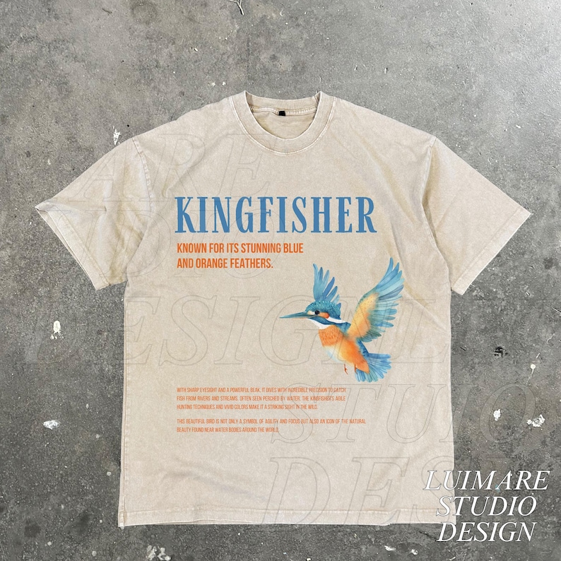 Puede incluir: Una camiseta beige con un dise&ntilde;o gr&aacute;fico de un mart&iacute;n pescador con plumas azules y naranjas. El texto "KINGFISHER" est&aacute; impreso en letras azules sobre el p&aacute;jaro. El texto "KNOWN FOR ITS STUNNING BLUE AND ORANGE FEATHERS." est&aacute; impreso en letras naranjas debajo del p&aacute;jaro.