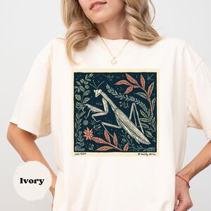 May include: Ivory-colored t-shirt featuring a detailed illustration of a praying mantis within a square frame. The artwork includes botanical elements in shades of green, orange, and cream against a dark blue background. The text "Semp 16014" and "@Healthy daring" are visible.