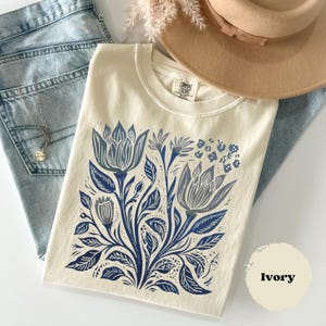 May include: Ivory-colored t-shirt with a blue floral design. The design features stylized flowers and leaves. The shirt is made of a soft material and has a crew neck. The tag reads "Comfort Colors".