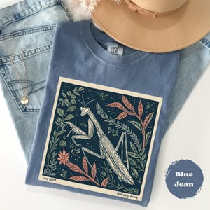 May include: A blue t-shirt with a graphic of a praying mantis surrounded by leaves and flowers. The shirt is folded on top of a pair of blue jeans, with a gold necklace and a tan hat in the background. The text "Blue Jean" is on the bottom right.