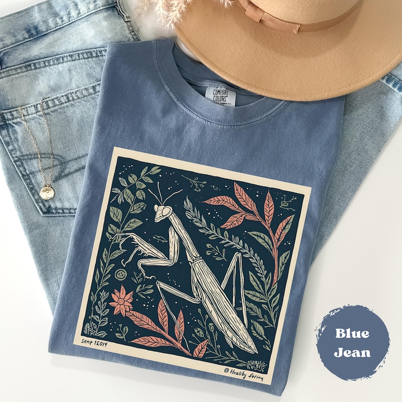 May include: A blue t-shirt with a graphic of a praying mantis surrounded by leaves and flowers. The shirt is folded on top of a pair of blue jeans, with a gold necklace and a tan hat in the background. The text "Blue Jean" is on the bottom right.
