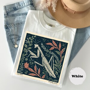 May include: White t-shirt featuring a detailed illustration of a praying mantis surrounded by foliage in shades of green, peach, and cream. The design is set against a dark blue background. The text "Healthy Faring" is visible.