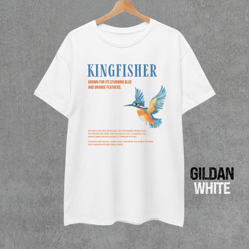Puede incluir: Camiseta blanca con un gr&aacute;fico de un mart&iacute;n pescador azul y naranja. El texto "KINGFISHER" est&aacute; impreso en negro sobre el p&aacute;jaro. El texto "KNOWN FOR ITS STUNNING BLUE AND ORANGE FEATHERS." est&aacute; impreso en naranja debajo del p&aacute;jaro. El texto "GILDAN WHITE" est&aacute; impreso en negro en la esquina inferior derecha de la camiseta.