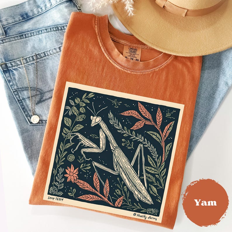 May include: An orange t-shirt with a graphic of a praying mantis surrounded by leaves and flowers. The shirt is folded on top of blue jeans, with a tan hat in the background. The shirt has the text "Comfort Colors" and "XL TG - EG".