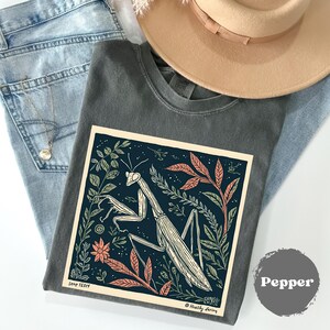 May include: A dark gray t-shirt with a large graphic of a praying mantis surrounded by leaves and flowers. The graphic is in a cream-colored frame. The shirt is folded on top of blue jeans and a tan hat.