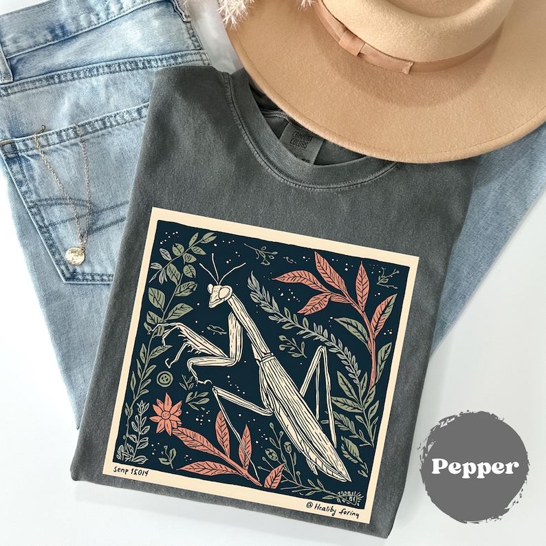 May include: A dark gray t-shirt with a large graphic of a praying mantis surrounded by leaves and flowers. The graphic is in a cream-colored frame. The shirt is folded on top of blue jeans and a tan hat.