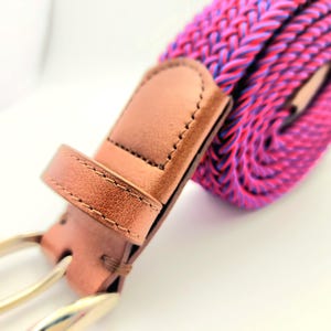 May include: Close-up of a brown leather belt with a silver buckle. The belt features a woven, elastic band in shades of pink, purple, and blue. The leather has visible stitching, and the buckle is a classic design.