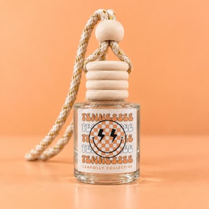 May include: A clear glass car air freshener bottle with a wooden cap and a braided rope hanger. The label features a checkered smiley face with lightning bolt eyes, the word TENNESSEE repeated, and the text LEAPOLLY COLLECTIVE at the bottom.