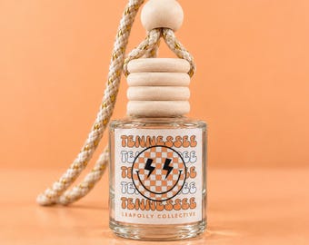 May include: A clear glass car air freshener bottle with a wooden cap and a braided rope hanger. The label features a checkered smiley face with lightning bolt eyes, the word TENNESSEE repeated, and the text LEAPOLLY COLLECTIVE at the bottom.