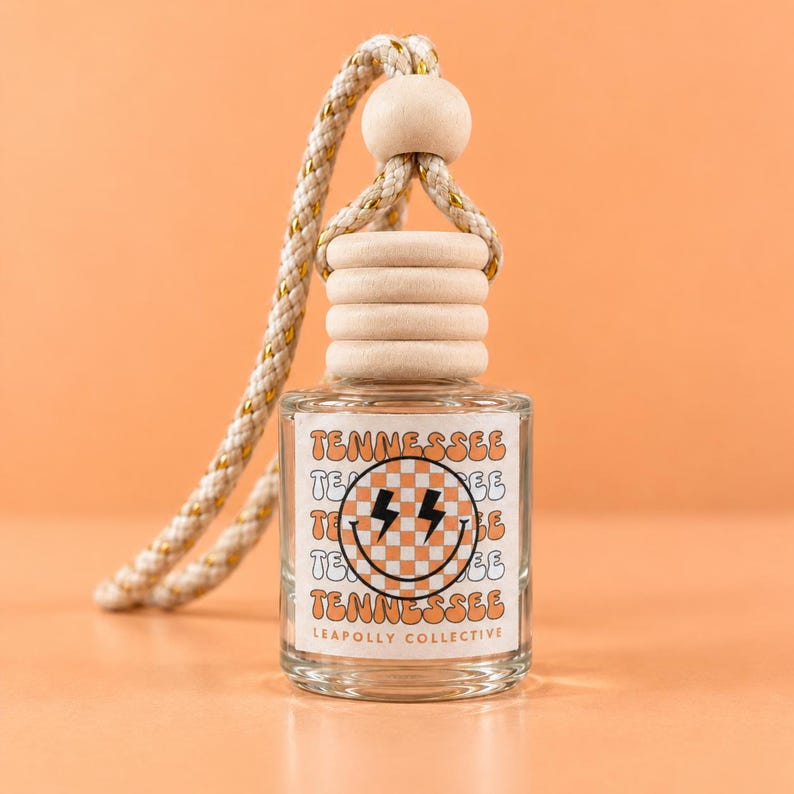 May include: A clear glass car air freshener bottle with a wooden cap and a braided rope hanger. The label features a checkered smiley face with lightning bolt eyes, the word TENNESSEE repeated, and the text LEAPOLLY COLLECTIVE at the bottom.