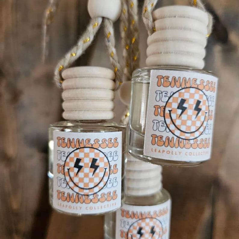May include: Three small clear glass car air freshener bottles with light wood screw-on caps, hanging from braided rope. Each bottle features a label with the text TENNESSEE and a checkered smiley face graphic with black lightning bolt eyes.
