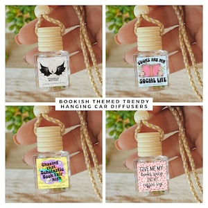 May include: Four hanging car diffusers with bookish themes. The diffusers are clear glass bottles with wooden stoppers and gold cords. The labels feature colorful illustrations and text, including "Wingspan Matters", "Books Are My Social Life", "Chasing That Scholastic Book Fair High", and "Give Me My Books Spicy And My Coffee Icy".