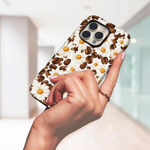 May include: A phone case with a design of white daisies with yellow centres and brown coffee beans on a white backdrop. The case has a glossy finish and a camera cut-out.
