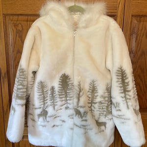 May include: Cream-coloured faux fur jacket with a hood and a forest scene design. The jacket features a zip closure and a detailed illustration of trees and deer in a winter landscape. The hood is trimmed with faux fur.