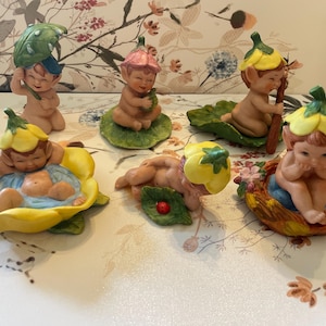 May include: A collection of ceramic fairy figurines. Each fairy is depicted in a unique pose, some with flower hats and others resting on leaves or in flower petals. The figurines have a pastel color palette, with details in green, yellow, pink, and blue.