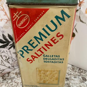 May include: Vintage Nabisco Premium Saltines tin. The rectangular tin has a light yellow background with red and teal lettering. The top and rim are teal. The tin reads "Premium Saltines" and "Galletas Delgaditas Tostaditas".