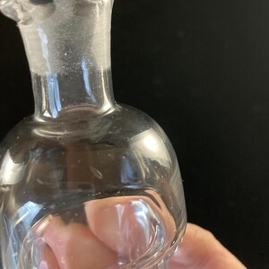 May include: A clear glass bottle with a rounded bottom and a narrow neck. The bottle has a slight lip around the opening.