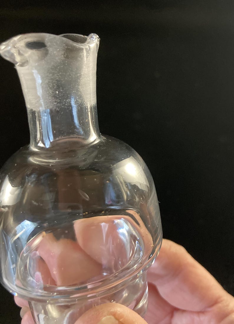 May include: A clear glass bottle with a rounded bottom and a narrow neck. The bottle has a slight lip around the opening.