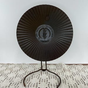 May include: A dark brown, circular metal object with a radial design, mounted on a wrought iron stand. The center of the object has embossed text that reads "PAT. PENDING" and "MADE IN USA". The stand has a circular base.