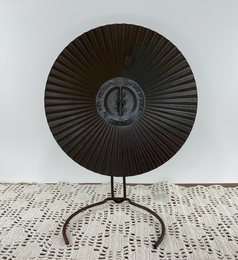 May include: A dark brown, circular metal object with a radial design, mounted on a wrought iron stand. The center of the object has embossed text that reads "PAT. PENDING" and "MADE IN USA". The stand has a circular base.
