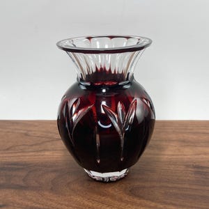 May include: A ruby red glass vase with a clear, fluted rim and a rounded body. The vase features a cut design of vertical leaf-like shapes. The vase sits on a wooden surface.