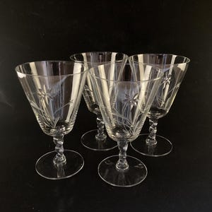 May include: Four clear, stemmed wine glasses with etched floral designs. The glasses have a flared bowl, a twisted stem, and a round base. The etched design features a star-like flower with delicate lines.