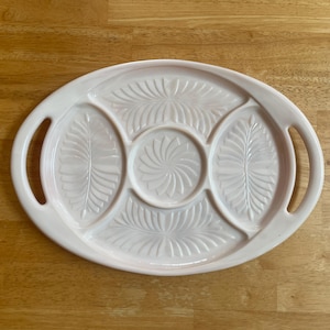 May include: An oval-shaped, pale pink serving tray featuring five distinct compartments. The tray includes two side handles and is decorated with embossed leaf and sunburst patterns pressed into the surface of each individual section.