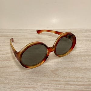 May include: Oval-shaped sunglasses with a tortoiseshell pattern. The frames are a mix of brown and amber hues, with dark gray lenses. The sunglasses are set against a light wood-grain surface.