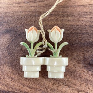 May include: A cream-colored tulip pendant with two flowers in matching pots, connected by a beige string. The tulip petals are tipped with peach, and the leaves are green. The pendant is on a wooden surface.