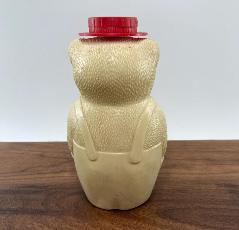 May include: A vintage bear-shaped bottle in a light beige color, wearing a red hat. The bear has textured details and is wearing overalls. The bottle is standing on a wooden surface.