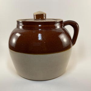 May include: A brown and white glazed ceramic crock with a lid and a handle. The crock is a traditional style with a rounded body and a wide mouth.