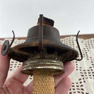 May include: Close-up of an antique oil lamp burner. The metal components are aged with a dark, rusty patina. A beige wick extends from the base, and the top features a charred wick. The lamp has a circular adjustment wheel and curved metal supports.