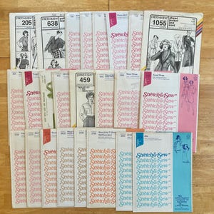 May include: A collection of vintage "Stretch & Sew" sewing patterns, featuring various garment designs. The patterns have a retro aesthetic, with illustrations of the finished garments and the pattern numbers visible. The color palette includes pink, blue, and white.