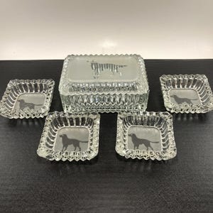 May include: A clear glass set featuring a rectangular box with a frosted top and a dog silhouette, along with four square ashtrays, each with a dog silhouette. The set has a textured, ridged design.
