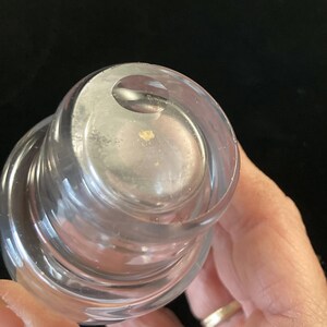 May include: Close-up of a clear glass bottle stopper with a layered design. The top of the stopper has a small, circular indentation.