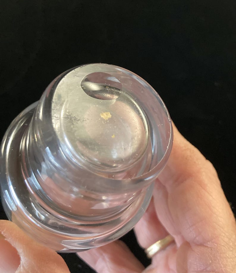 May include: Close-up of a clear glass bottle stopper with a layered design. The top of the stopper has a small, circular indentation.