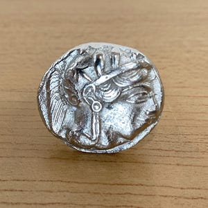 May include: A silver-colored coin featuring a detailed profile of a person with flowing hair and a laurel wreath. The coin has an aged, antique appearance, with visible texture and wear. The coin is round and appears to be made of metal.