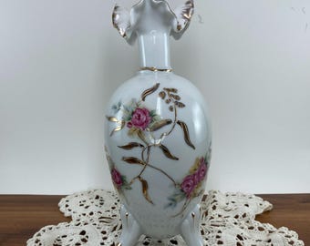 May include: A white porcelain vase with a ruffled top and three feet. The vase is decorated with pink roses, gold accents, and a delicate floral design. The vase is approximately 8 inches tall.