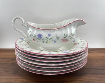 May include: A white ceramic gravy boat with a handle and a stack of matching plates. The gravy boat and plates feature a floral pattern with pink, blue, and yellow flowers. The rims of the plates are trimmed in pink.