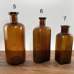 May include: Three antique amber glass bottles of varying sizes. The bottles have dark brown stoppers and are displayed on a wooden surface. The bottles are cylindrical, rectangular, and square-shaped.