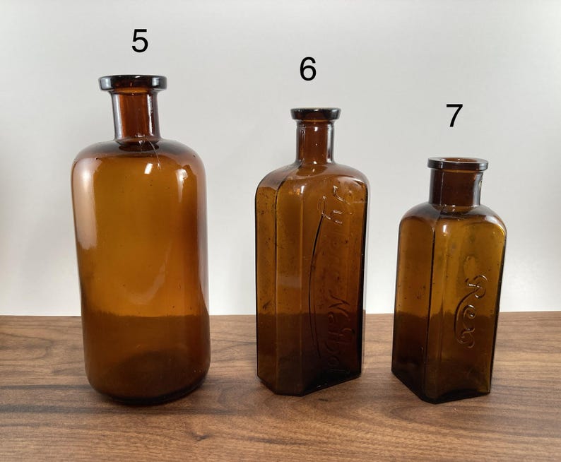 May include: Three antique amber glass bottles of varying sizes. The bottles have dark brown stoppers and are displayed on a wooden surface. The bottles are cylindrical, rectangular, and square-shaped.