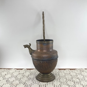 May include: A hammered copper vessel with a rounded body, a pedestal base, and a side spout. The vessel features a tall, vertical, twisted metal handle attached to the rim. The metal has a dark, aged patina with visible texture.