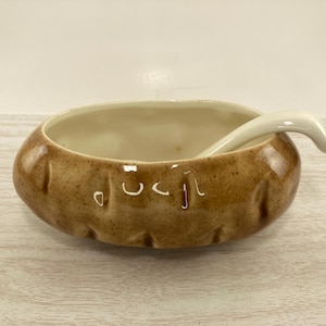 May include: A ceramic potato-shaped bowl with a matching spoon. The bowl is brown with darker brown speckles and has a light beige interior. The spoon is white and curved. The bowl has a rustic, handmade appearance.