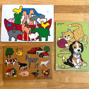 May include: Three wooden jigsaw puzzles featuring colorful illustrations. One puzzle depicts Noah's Ark with various animals, another shows a farm scene with livestock and buildings, and the third displays a cat and a dog playing with yarn.