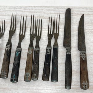 May include: A collection of antique silverware, including forks and knives. The forks have tines and wooden handles with decorative metal accents. The knives have blades and wooden handles. The silverware is arranged on a light-colored wooden surface.