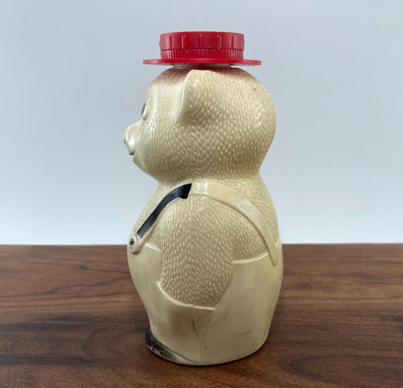 May include: A vintage pig-shaped bottle with a red cap. The bottle is beige with a textured surface and features a pair of cream-colored overalls with black straps. The pig has a small red hat on its head.