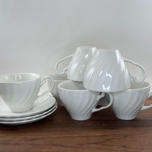 May include: A set of six white ceramic teacups and saucers. The cups have a spiral design and curved handles. The saucers are stacked, and one cup sits on top of the stack. The teacups and saucers are on a wooden surface.