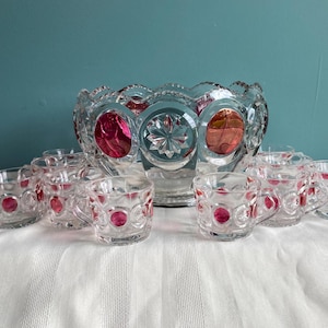 May include: A clear glass punch bowl and matching cups set. The bowl and cups feature a decorative pattern with red accents. The set is displayed on a white tablecloth against a teal background. The cups have small handles.