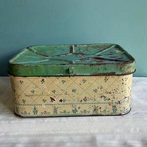May include: A rectangular metal storage tin with a distressed green lid and a cream-colored base. The base features a blue diamond grid pattern with small floral accents and a bottom border depicting windmills and figures in traditional clothing.